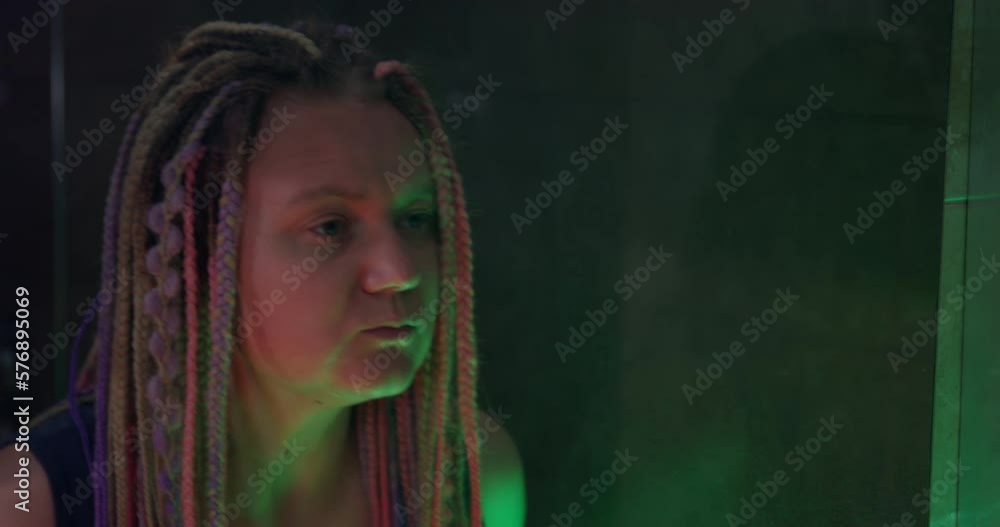 Stylish young woman with multicolored box braids hairstyle appears in the frame. She looks at herself in the mirror and explores her hair. Dim room is lit by colored lamps. Stylish young woman with multicolored box braids hairstyle appears in the frame. She looks at herself in the mirror and explores her hair. Dim room is lit by colored lamps.