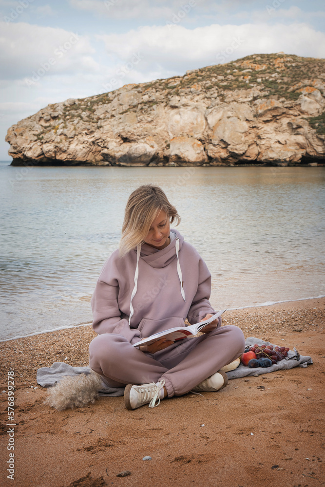 Portrait of a blonde girl with fair skin. A woman sits on the beach and reads a book, eats and relaxes, enjoys the moment. Self love, meditation on the ocean, rocks. High quality photo
