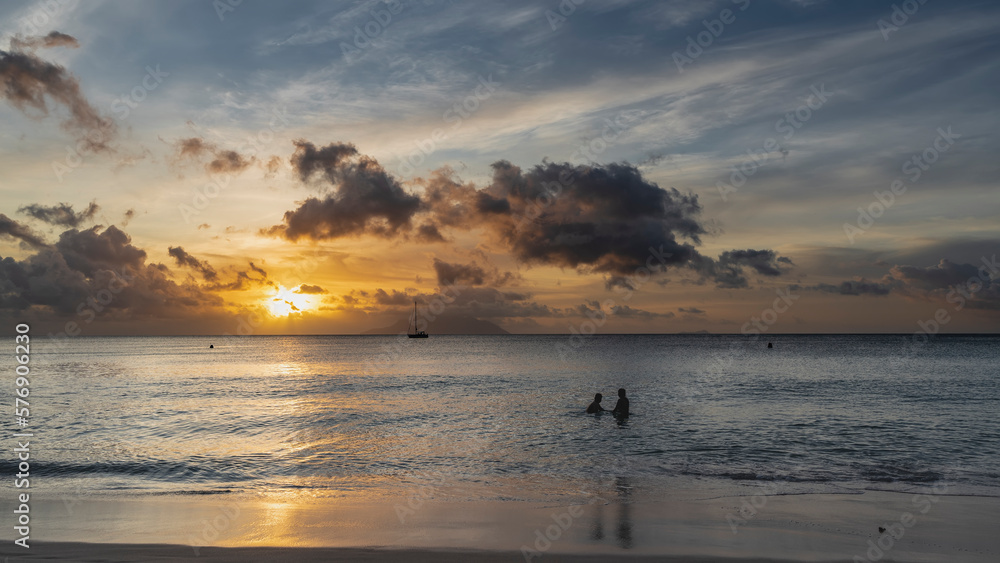 Sunset on a tropical island. The sun shines through clouds colored pink and purple. The yacht is on the horizon. Silhouettes of two people are visible in the ocean. Reflection on wet shiny sand. 
