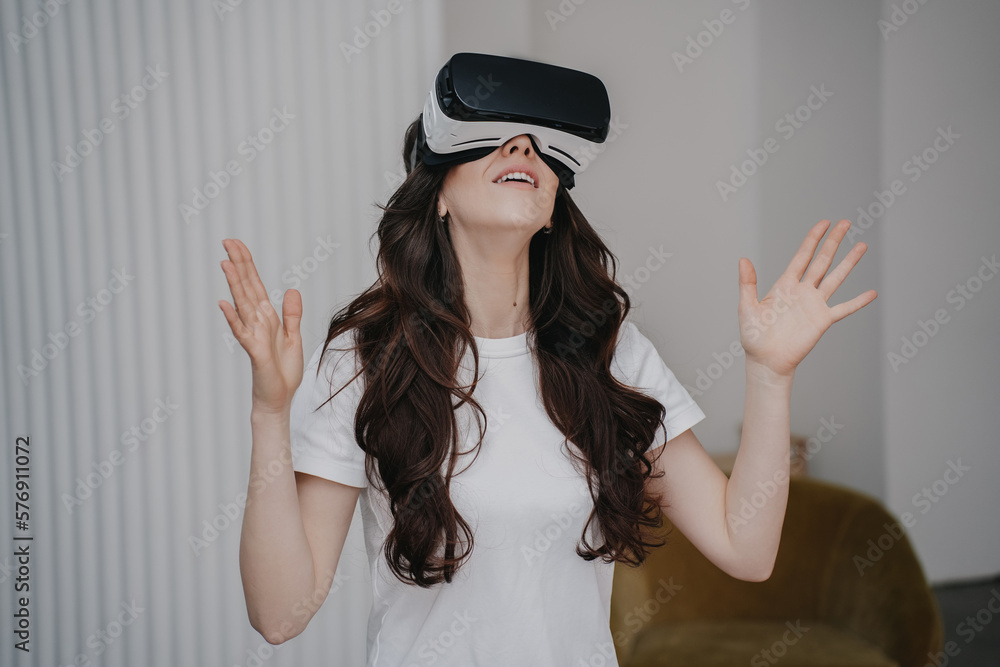 Pretty brunette with loose wavy hair in white t-shirt using virtual headset at home, playing at weekend. Young adult hispanic woman spending time with virtual reality glasses over blurry interior. Pretty brunette with loose wavy hair in white t-shirt using virtual headset at home, playing at weekend. Young adult hispanic woman spending time with virtual reality glasses over blurry interior.