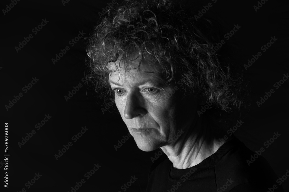 Black and white portrait of a middle aged woman with a neutral expression, looking downward.  Low key background.