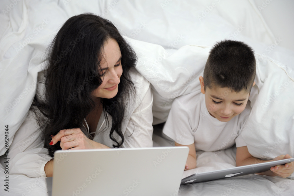 work home online woman under blanket working on laptop beside son playing games on tablet sick at home get well family isolation quarantine white sheet duvet black hair mom and son work home online woman under blanket working on laptop beside son playing games on tablet sick at home get well family isolation quarantine white sheet duvet black hair mom and son