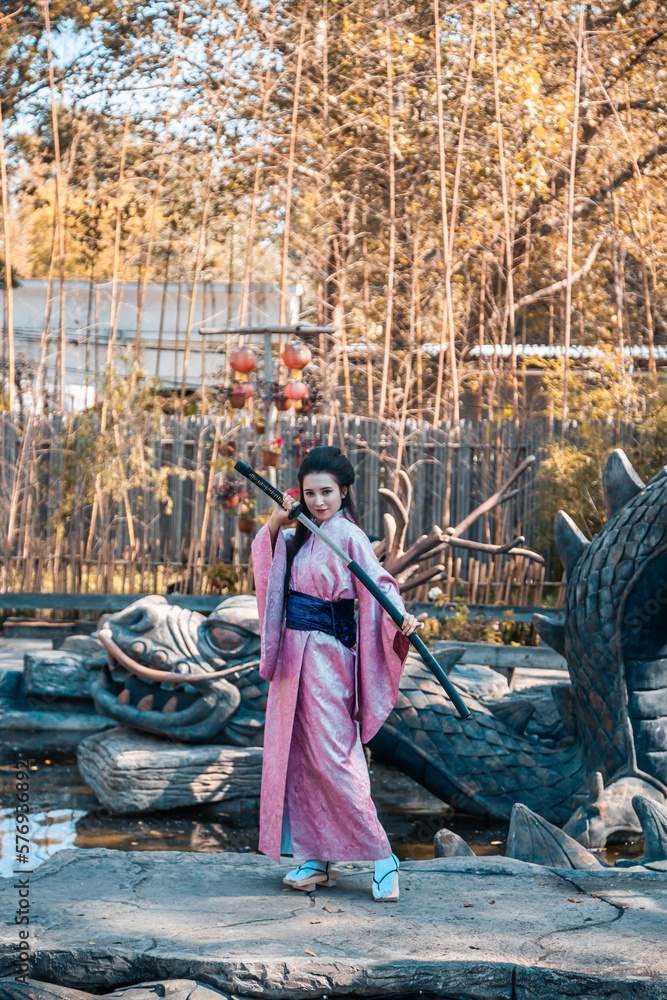 Japanese geisha in a traditional kimano with a fan and armed with a katana sword in a beautiful garden. A girl from medieval Asia. Reconstruction of cultural heritage. Culture in Japan.