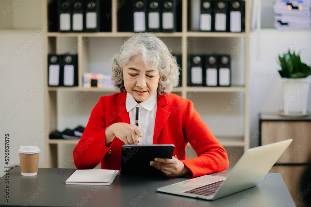 Old beautiful woman using laptop and tablet while sitting at her working place. Concentrated Old beautiful woman using laptop and tablet while sitting at her working place. Concentrated