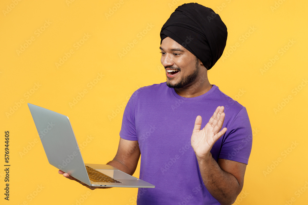 Devotee Sikh Indian IT man ties his traditional turban dastar wear purple t-shirt work hold use point laptop pc computer waving hand get video call isolated on plain yellow background studio portrait. Devotee Sikh Indian IT man ties his traditional turban dastar wear purple t-shirt work hold use point laptop pc computer waving hand get video call isolated on plain yellow background studio portrait.