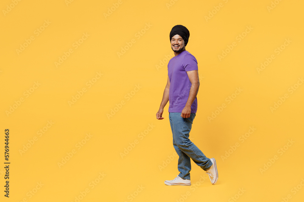 Full body fun side view smiling cheerful devotee Sikh Indian man ties his traditional turban dastar wear purple t-shirt walking going look camera isolated on plain yellow background studio portrait. Full body fun side view smiling cheerful devotee Sikh Indian man ties his traditional turban dastar wear purple t-shirt walking going look camera isolated on plain yellow background studio portrait.