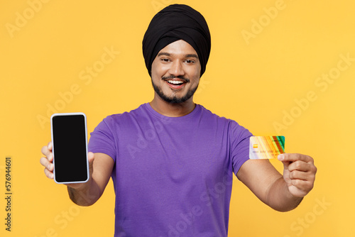 Fun devotee Sikh Indian man ties his traditional turban dastar wear purple t-shirt use blank screen mobile cell phone credit bank card shopping online booking tour isolated on plain yellow background. photo