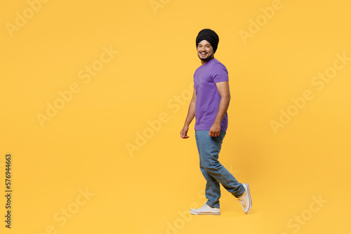 Full body fun side view smiling cheerful devotee Sikh Indian man ties his traditional turban dastar wear purple t-shirt walking going look camera isolated on plain yellow background studio portrait.
