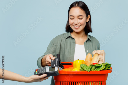 Young woman in casual clothes hold red basket with food products hold bank payment terminal process acquire credit card isolated on plain blue background studio portrait. Delivery service from shop.