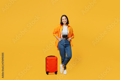 Young smiling woman wear summer casual clothes stand near suitcase baggage isolated on plain yellow background. Tourist travel abroad in free spare time rest getaway. Air flight trip journey concept.