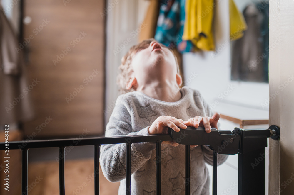 Front view of a little boy standing on the other side of the child safety gate. A little boy is holding on to the top of the security gate and looking at the camera from above. Front view of a little boy standing on the other side of the child safety gate. A little boy is holding on to the top of the security gate and looking at the camera from above.