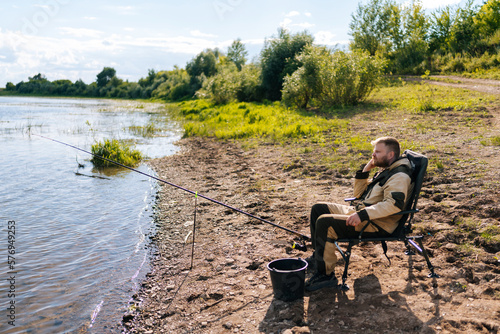 Side view of pensive bearded fisherman sitting on river bank on travel chair waiting for catch looking around. Patient fisher man fishing outdoors in river enjoying weekend leisure.