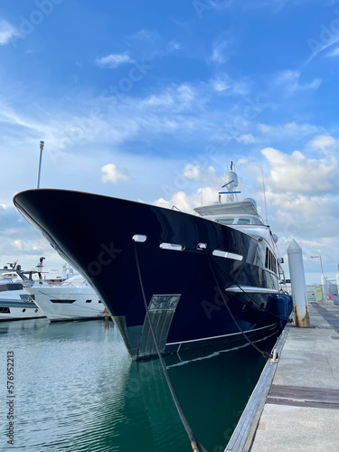 Ship in the port. Front of a large luxury pleasure yacht in the foreground. Yachts moored to the pier in the background. Vertical frame. Boat's marina. Beautiful blue sly with clouds. Yacht close-up