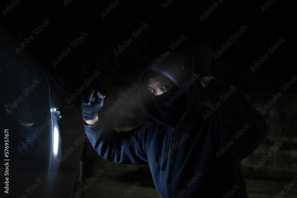 Young thief in black is stealing a car at night. He carries a flashlight and a screwdriver for car theft.