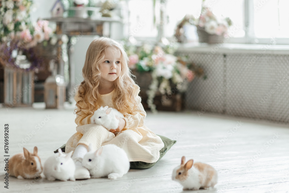 A little smiling girl in a yellow dress is playing on the floor in a room with rabbits. A baby and a rabbit. The living room is decorated for Easter. A little smiling girl in a yellow dress is playing on the floor in a room with rabbits. A baby and a rabbit. The living room is decorated for Easter.