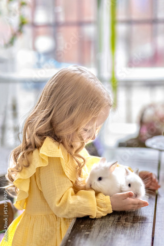 A little blonde girl in a yellow dress is sitting at a festive Easter table with rabbits.A baby and a rabbit. The concept of Easter.Easter at the table. The Christian tradition.