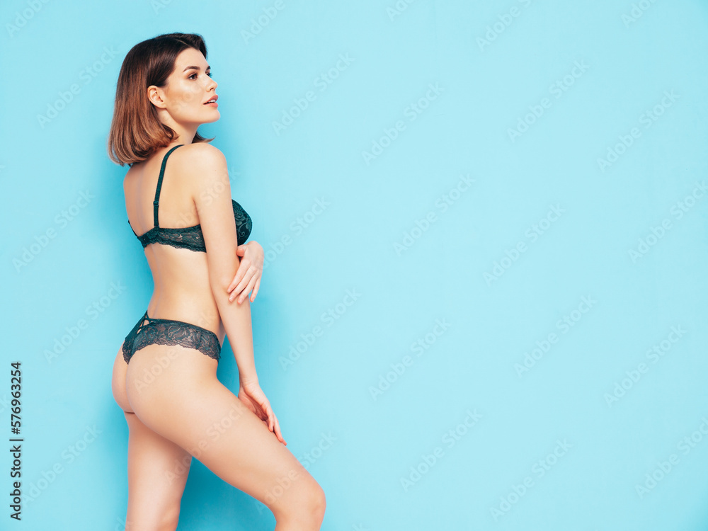 Naklejka premium Portrait of young beautiful sexy woman in green lace lingerie. Carefree smiling model wearing pure underwear. Hot brunette posing near blue wall in studio. Perfect body. Slim and fit. Isolated