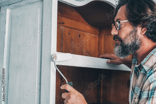 Close up of mature man painting and restoring an old antique wooden cabinet at home with roller and white paint. Recycling and upcycling oldies. People working in leisure hobby activity alone photo