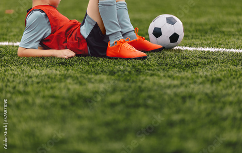 Young soccer boy with the ball lying and resting at soccer grass field at pitch sideline. Child at break time during soccer football training session