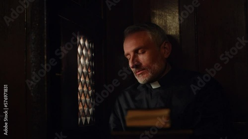 The gray-haired pastor opens the window in the confessional and listens attentively to the parishioner. A priest in a black robe listens to confession. photo