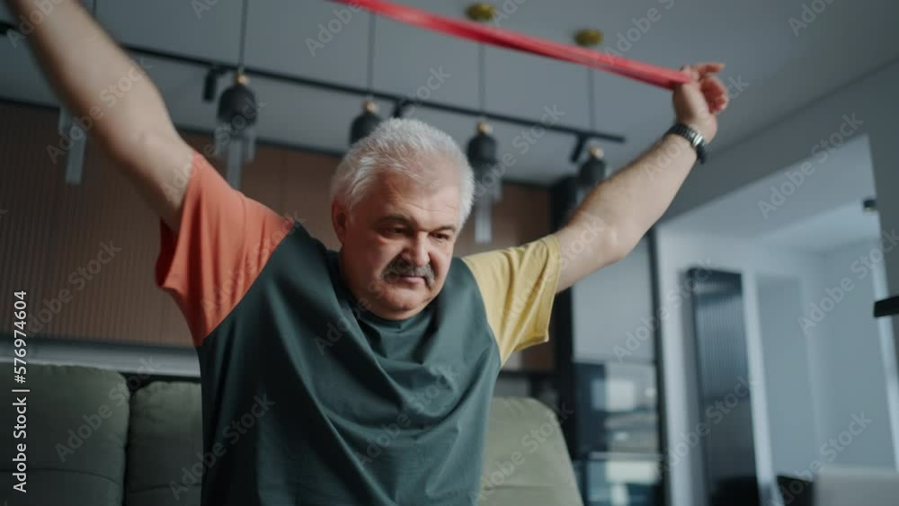 Aged Man Doing Exercise For Spinal Health, Using Elastic Band, Watching Online Tutorial Aged Man Doing Exercise For Spinal Health, Using Elastic Band, Watching Online Tutorial