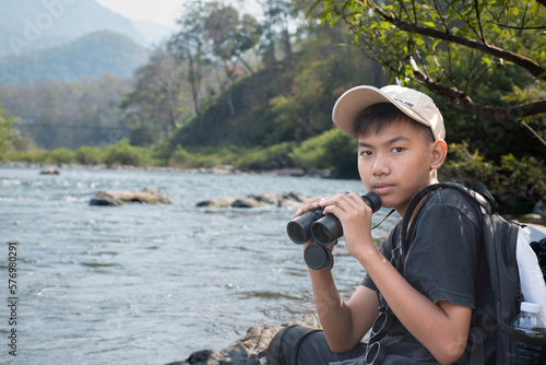 Asian boy sitting by river bank and using binoculars to watch birds on the trees in local national park during summer camp, idea for learning creatures and wildlife animals outside the classroom.