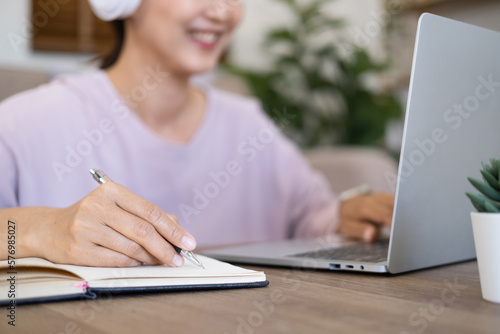 Close up hand - student wear wireless headphone study online with skype teacher, happy young woman learn language listen lecture watch webinar write notes look at laptop in home , distance education