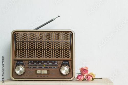 Against the background of the white wall on the shelf, there is a brown vintage retro of brown radio, next to dry wilting roses, copy the space.