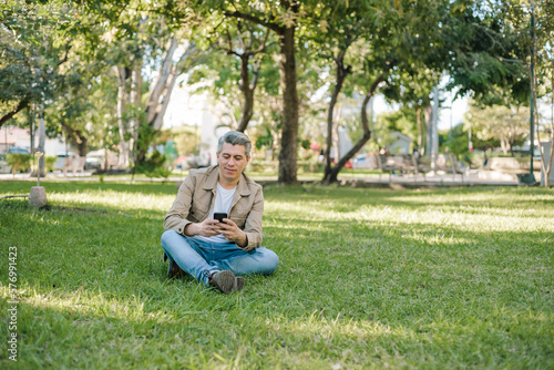 Gray-haired man looking at his cell phone sitting on the grass in a park.