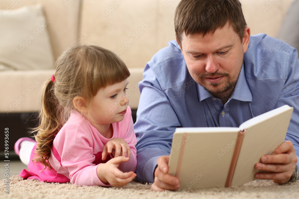 Father is reading book to daughter lying on floor in bedroom