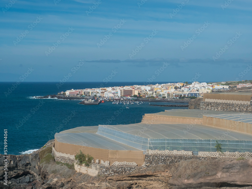 distant aerial view of Port of Los Cristianos with fishermans boat and sailing ships and city building at background. Tenerife, Canary Islands, Spain, Sunny day