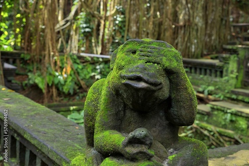 Close-up of a moss-covered stone figure in the shape of a frog in the middle of a sun-drenched rainforest  with a temple complex diffused in the background.