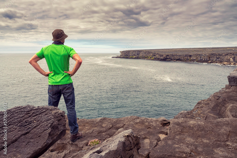 Male tourist in green shirt standing on edge of a cliff looking at amazing scenery. Aran Island, Ireland. Travel and tourism. Rough Irish nature landscape.
