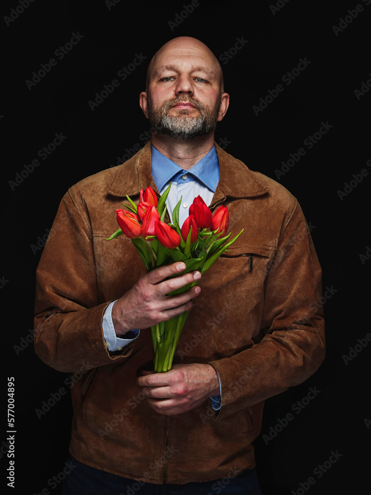 Bald man holding bouquet of red tulips. Male model in his 40s with grey beard and wearing brown suede leather jacket with flowers. Dark background. Expression of emotion towards his woman. Bald man holding bouquet of red tulips. Male model in his 40s with grey beard and wearing brown suede leather jacket with flowers. Dark background. Expression of emotion towards his woman.
