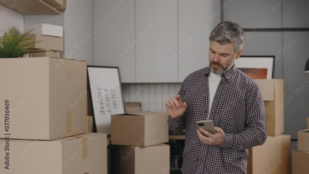 Middle Aged Man Is Using Smartphone While Relocation Standing in New Apartment With Cardboard Boxes on the New Kitchen Background. Male Person Calculating Costs on Mobile. Middle Aged Man Is Using Smartphone While Relocation Standing in New Apartment With Cardboard Boxes on the New Kitchen Background. Male Person Calculating Costs on Mobile.