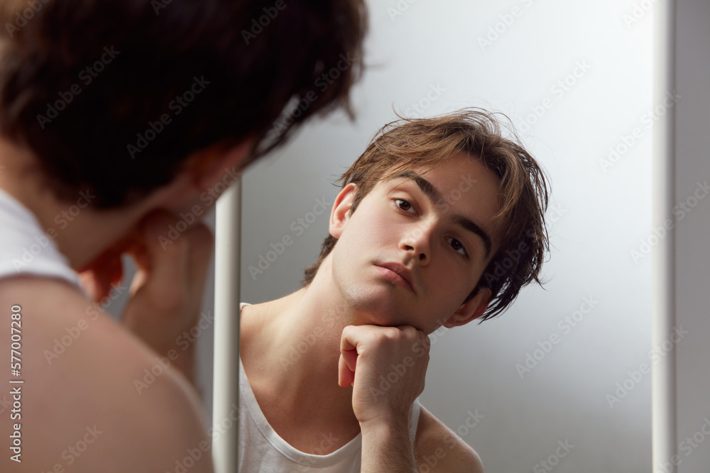 Portrait of young man looking with well-kept skin, shaved face looking in mirror against grey studio background. Concept of men's health, body and skin care, hygiene and male cosmetology Portrait of young man looking with well-kept skin, shaved face looking in mirror against grey studio background. Concept of men's health, body and skin care, hygiene and male cosmetology