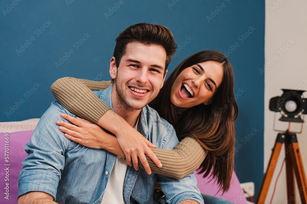 Fototapeta premium Close up portrait of happy caucasian young couple hugging and smiling together in a couch at home. Beautiful brunette wife embracing her husband resting in a sofa indoors. High quality photo