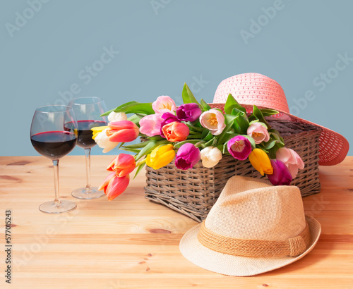 Colorful fresh tulips in wicker basket, hats and glasses of red wine on wooden table.