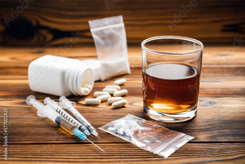 Glass with alcohol drink, whisky or brandy, white pills, syringe with a drugs dose, narcotic in transparent bag on a wooden table. Concept of addiction and bad habits