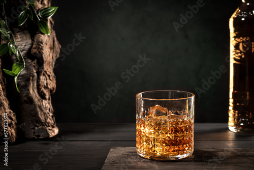 Glass of whisky with ice and bottle on wooden table on dark background with copy space.