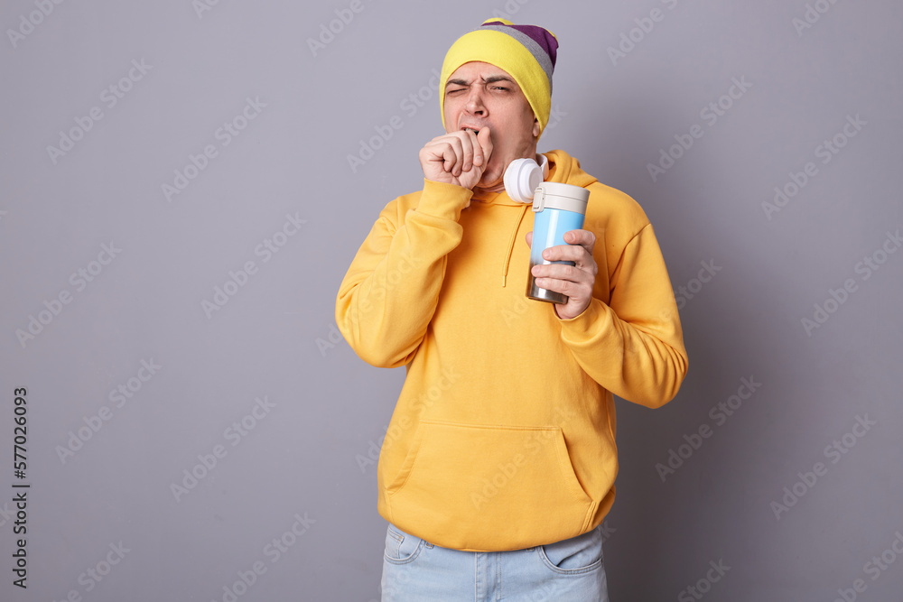 Photo of sleepy Caucasian man wearing beanie hat and yellow hoodie posing against gray wall, standing with thermos in hands, drinking coffee, needs energy, yawning, covering mouth. Photo of sleepy Caucasian man wearing beanie hat and yellow hoodie posing against gray wall, standing with thermos in hands, drinking coffee, needs energy, yawning, covering mouth.
