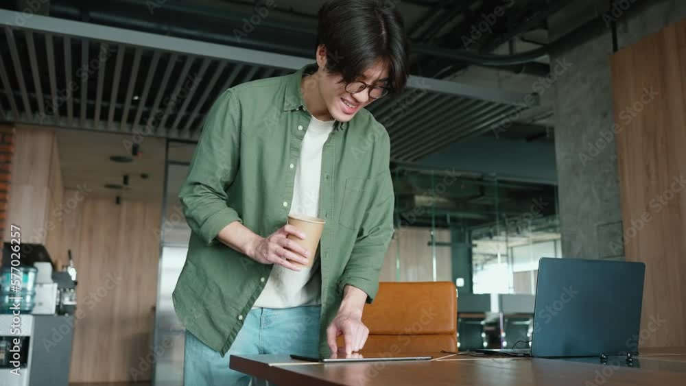 Happy brunet Asian man working on a digital tablet with cup of coffee in the office Happy brunet Asian man working on a digital tablet with cup of coffee in the office