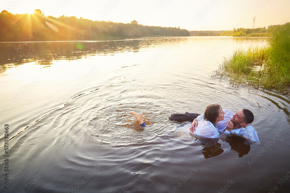 Fototapeta premium A beautiful adult couple having hugs and fun on nature in the water of a river or lake in the summer evening at sunset. A guy and a girl swim and relax outdoors in clothes in white shirts and jeans