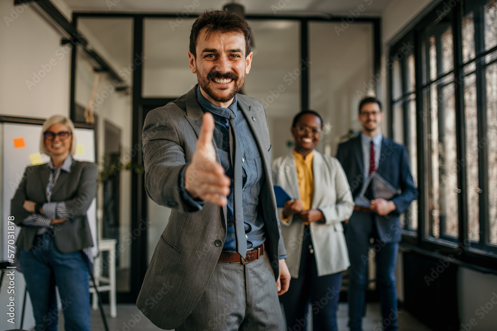 Portrait of handsome smiling businessman with his colleagues. Multi-ethnic group of business persons standing in modern office. The team leader approaches the hand for a handshake toward the camera. Portrait of handsome smiling businessman with his colleagues. Multi-ethnic group of business persons standing in modern office. The team leader approaches the hand for a handshake toward the camera.