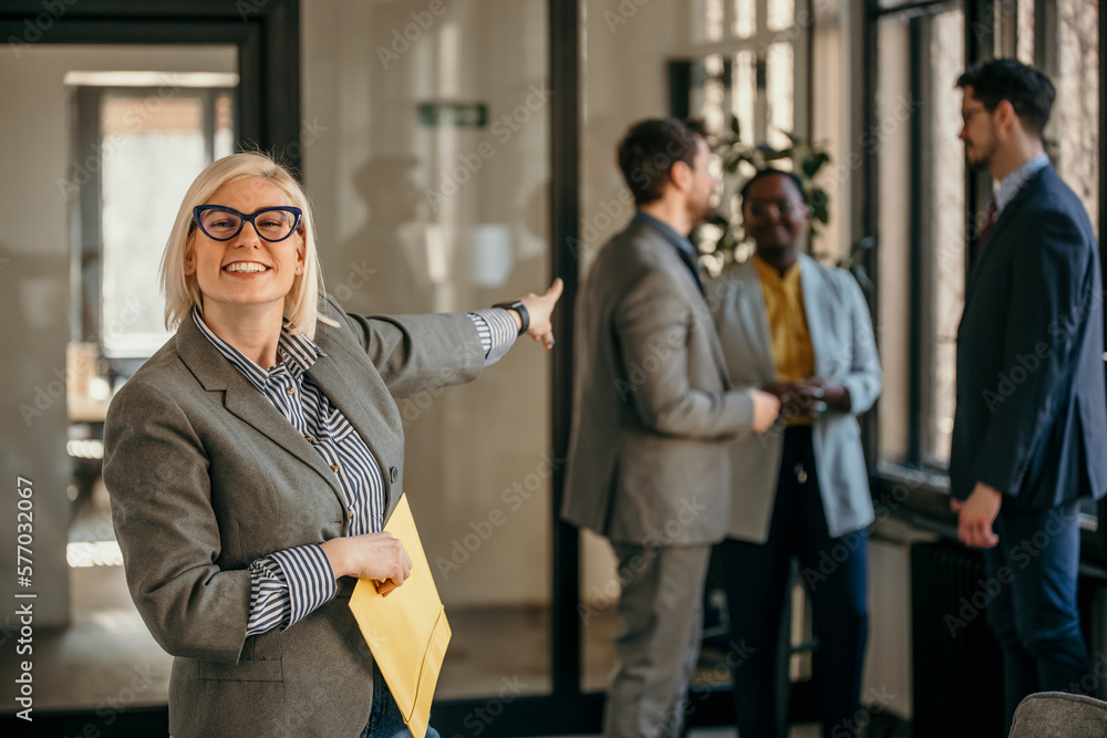 Shot of an attractive young blond businesswoman standing in the office holding files while pointing at the team and her colleagues standing behind her. Shot of an attractive young blond businesswoman standing in the office holding files while pointing at the team and her colleagues standing behind her.