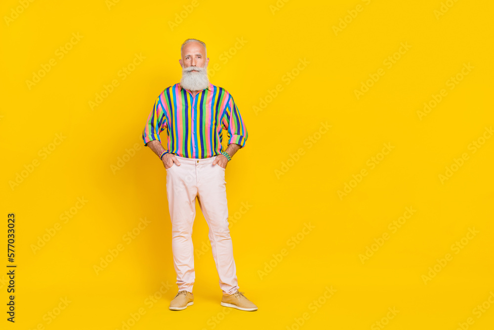 Fototapeta premium Photo of handsome retired man with white beard dressed striped shirt white trousers arms in pockets isolated on yellow color background