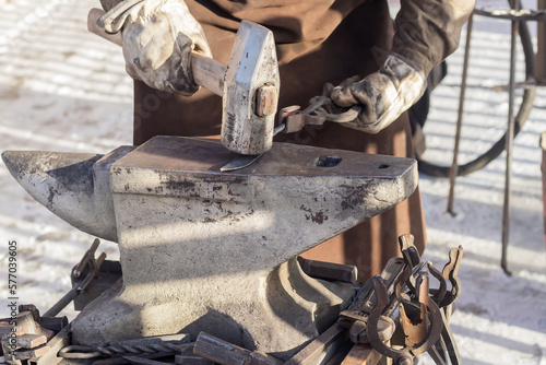 A blacksmith with a heavy hammer in his hand is working on an old anvil outdoors in the courtyard of the workshop.