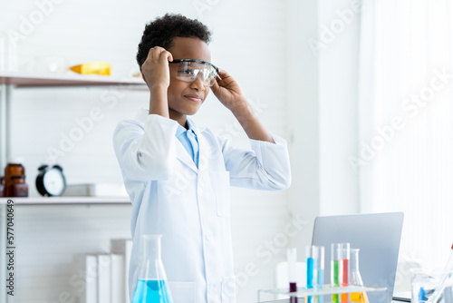 In chemistry classroom with many laboratory tools. A young African boy in white lab coat wearing safety glasses and smile about his new idea for next experiment with many colorful test tubes on table.