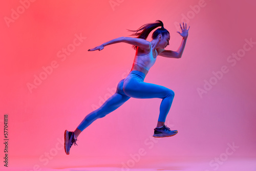 Dynamic portrait of young active girl, athlete, runner in motion, training over pink studio background in neon light. Concept of sportive lifestyle, health, endurance, action and motion. Ad