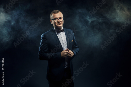 Handsome confident businessman in glasses wearing suit with bow tie standing over dark background
